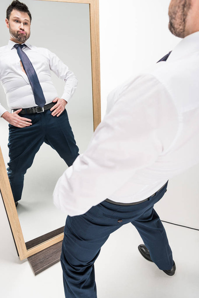 Übergewichtiger Mann in formeller Kleidung sieht Spiegelung im Spiegel und macht Gesichtsausdruck auf Weiß - Foto, Bild