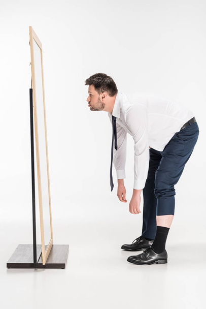 Übergewichtiger Mann in formaler Kleidung beugt sich vor Spiegel auf weiß - Foto, Bild