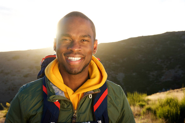 Nahaufnahme Porträt eines hübschen jungen afrikanisch-amerikanischen Mannes mit Rucksack, der in der Natur mit Sonnenuntergang im Hintergrund posiert - Foto, Bild