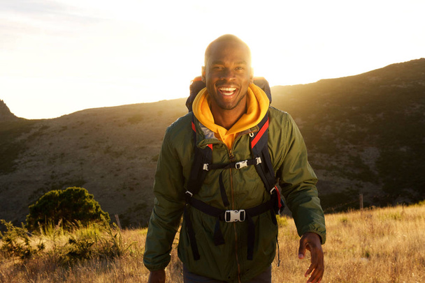 Portrait of happy african american man walking in with backpack in mountains and sunset in background - Photo, Image