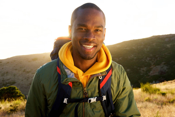 Close-up portret van knappe jonge Afro-Amerikaanse man met rugzak poseren in de natuur zonsopgang in de achtergrond - Foto, afbeelding