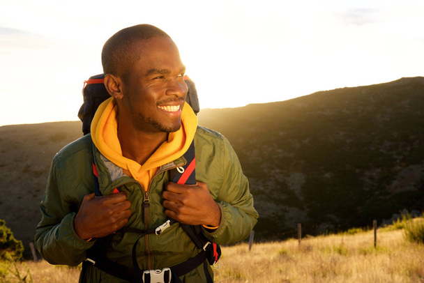 Close up portrait of handsome young african american man with backpack smiling with sunset in background - Photo, Image