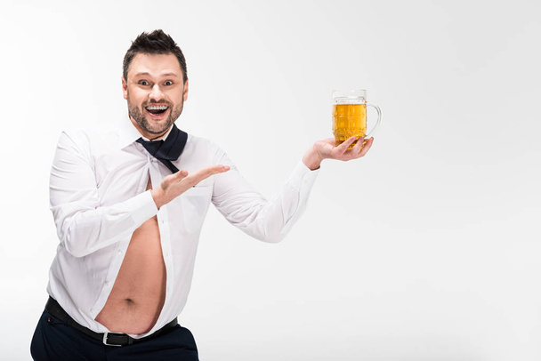 happy overweight man in tight shirt holding glass of beer and gesturing with hand isolated on white with copy space - Fotó, kép