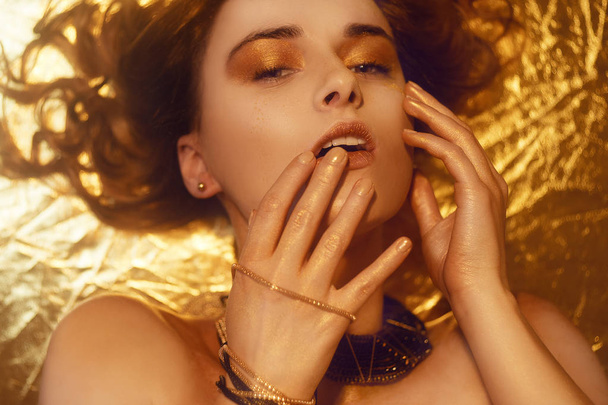 Gold Fashion Makeup, Art Beauty Face and Lips Make Up in Golden brocade. Fashion golden skin Woman face portrait. Gold jewellery, jewelry, accessories, earrings bracelet. Golden shadows. Girl lies. - 写真・画像
