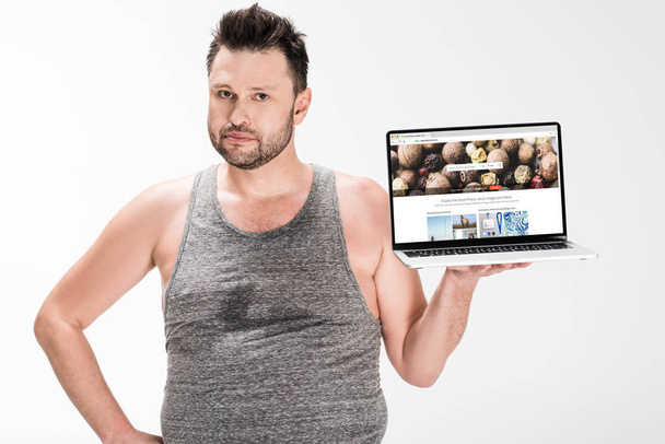 overweight man looking at camera and holding laptop with depositphotos website on screen isolated on white - Foto, imagen