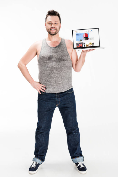 smiling overweight man looking at camera and presenting laptop with ebay website on screen isolated on white - Foto, afbeelding