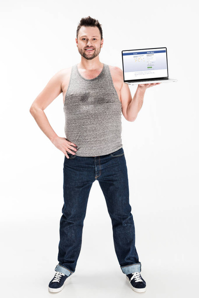 uomo in sovrappeso sorridente guardando la fotocamera e presentando laptop con facebook sito web sullo schermo isolato su bianco
 - Foto, immagini