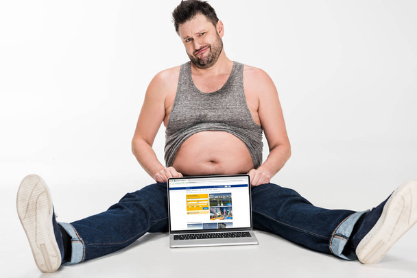 skeptical overweight man making facial expression and sitting with laptop with booking website on screen isolated on white - Foto, Bild