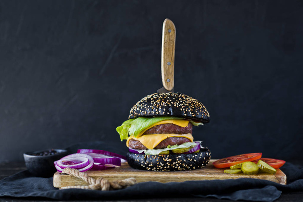schwarzer Hamburger auf einem hölzernen Schneidebrett. Seitenansicht, schwarzer Hintergrund, Platz für Text. - Foto, Bild