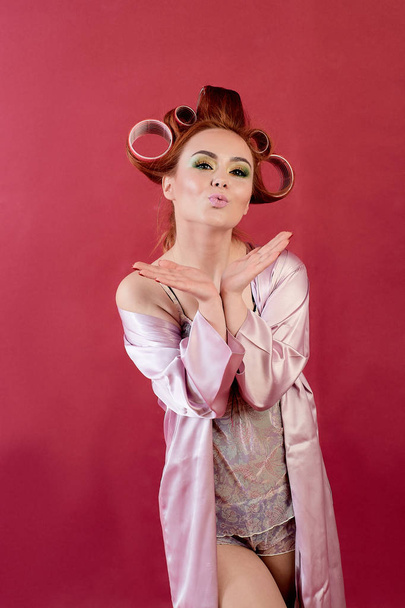 Young redhead woman with hair curlers dressed in neglige, peignoir, dressing gown posing on a burgundy background. - Foto, Bild