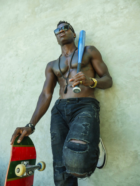 giovane attraente e bello nero afroamericano uomo con vestibilità corpo muscolare e sei pack in possesso di mazza da baseball e skate board posa cool urbano cazzuto
 - Foto, immagini