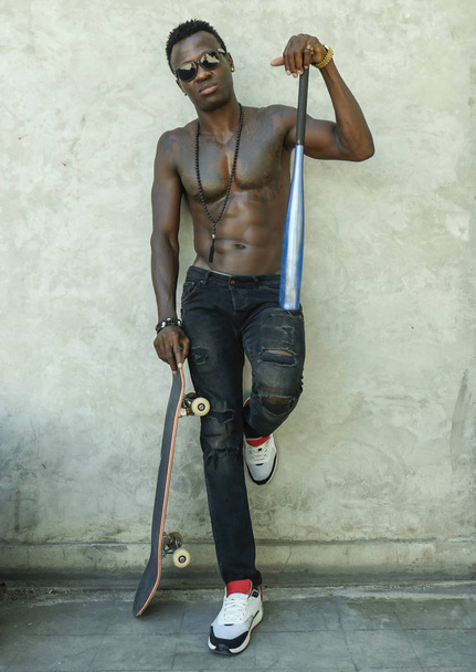 young attractive and handsome black African American man with fit muscular body and six pack holding baseball bat and skate board posing cool urban badass - Foto, Bild