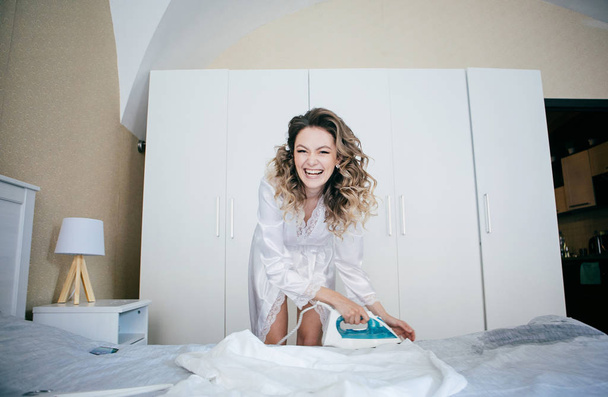 girl in a white silk dressing gown stroking an iron on the bed - Foto, Bild