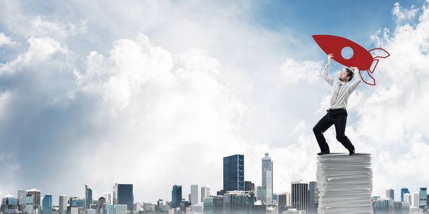 Confident young businessman in suit standing on paper columns and launching big drawn rocket with modern cityscape view on background - Photo, Image