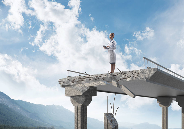 Conceptual image of woman doctor in white sterile suit standing on edge of broken bridge with landscape view on background. Medical industry concept - Photo, Image