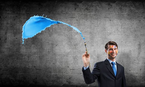 Successful businessman in black suit gesturing and smiling while standing with blue coloured splash against gray concrete wall on background. - Photo, Image