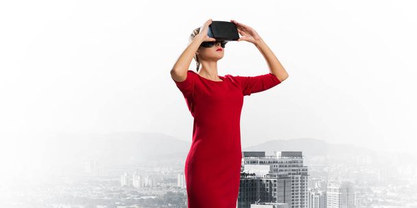 背景に都市景観のビューに立っている間、バーチャルリアリティヘッドセットを使用して赤いドレスを着た若い女性の概念的なイメージ。最新のテクノロジー - 写真・画像