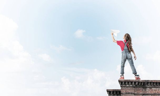 Χαριτωμένο χαρούμενο κορίτσι στο κτίριο κορυφή παίζει με χάρτινο αεροπλάνο - Φωτογραφία, εικόνα