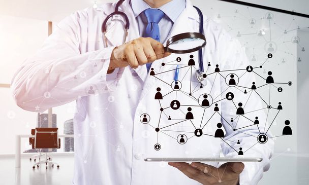 professionelle und junge Ärztin in weißer medizinischer Uniform schaut durch eine Lupe auf die soziale Netzwerkverbindung, während sie im hellen Bürogebäude steht - Foto, Bild