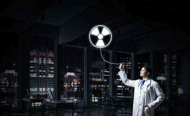 Young medical industry employee interracting with glowing radioactive symbol while standing against night cityscape view on background. - Photo, Image