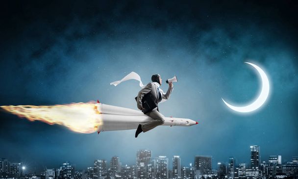 Conceptuele afbeelding van jonge zakenman in pak vliegen op raket met stadsgezicht en blauw donkere nachthemel met maan op achtergrond. - Foto, afbeelding