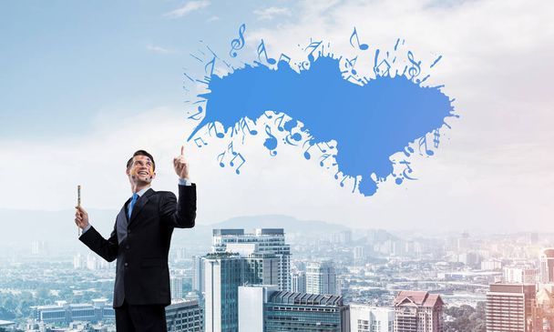 Horizontal shot of cheerful and young businessman in black suit gesturing and smiling while standing with blue coloured musical splash against cityscape view on background. - Photo, Image