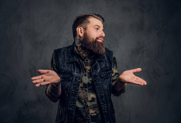 A stylish bearded guy with tattooed hands in military shirt and denim vest posing with a puzzled look. Studio photo against dark wall - Photo, Image