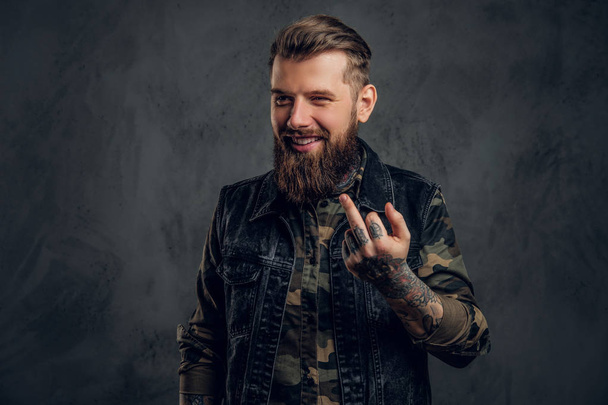 Сумасшедший бородатый мужчина в военной рубашке и джинсовом жилете с татуированными руками, показывающими ебаный знак. Студийное фото на фоне темной стены
 - Фото, изображение