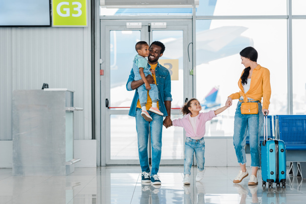 glimlachende Afro-Amerikaanse familie lopen met bagage langs wachtzaal in de luchthaven - Foto, afbeelding
