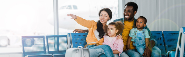 plano panorámico de la sonriente familia afroamericana con equipaje y niños sentados en el aeropuerto mientras la madre señala con el dedo de distancia
 - Foto, imagen
