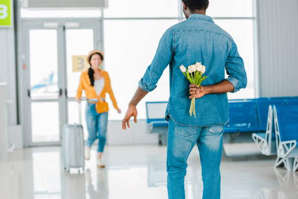 foyer sélectif de l'homme afro-américain marche vers petite amie heureuse avec valise tout en cachant des tulipes derrière son dos à l'aéroport
 - Photo, image