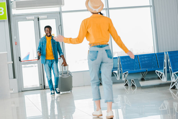 Selektywny fokus szczęśliwy African American Man z bagażem chodzenie do dziewczyny na lotnisku - Zdjęcie, obraz