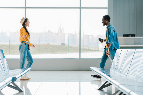 vista lateral do homem americano africano feliz andando em direção a namorada sorridente no aeroporto
 - Foto, Imagem