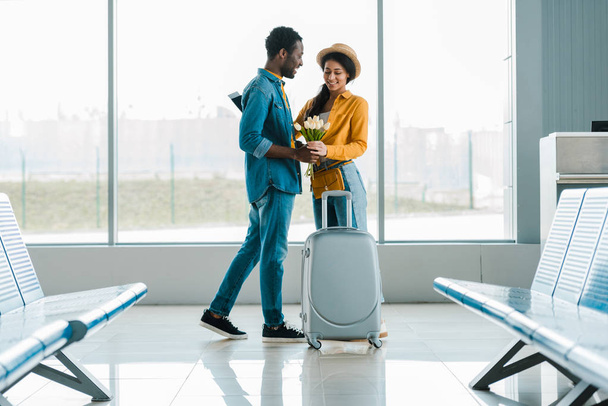 smiling african american man gifting bouquet of tulips to girlfriend with suitcase in airport - Photo, Image