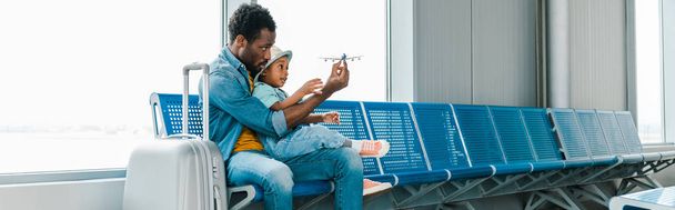 plano panorámico de padre e hijo afroamericano sentado con maleta en el aeropuerto y jugando con avión de juguete
 - Foto, imagen