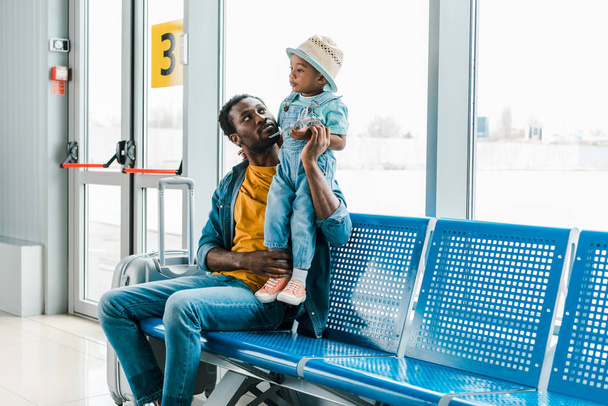 Afro-Amerikaanse vader zit met koffer in de wachtzaal in de luchthaven en kijken naar Son - Foto, afbeelding