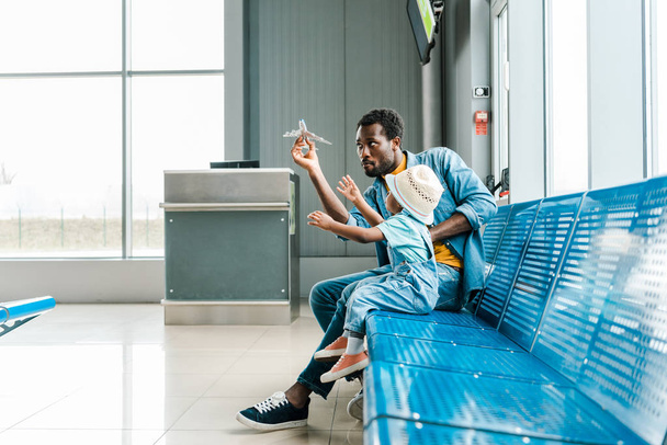 centrado afroamericano padre e hijo sentado en la sala de espera en el aeropuerto y jugar con avión de juguete
  - Foto, imagen