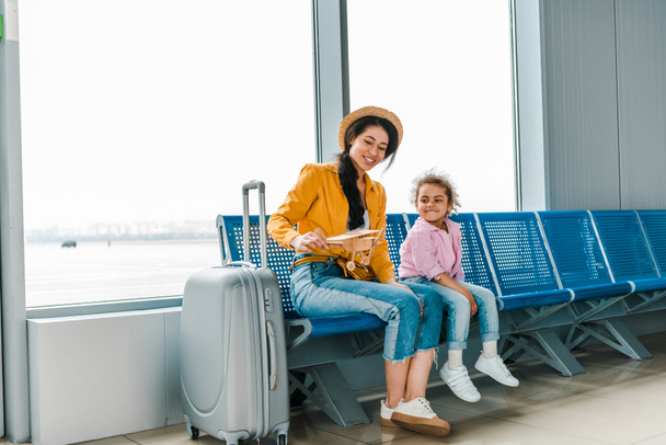 smiling african american mother and daughter sitting in airport with suitcase and wooden plane model  - Фото, изображение