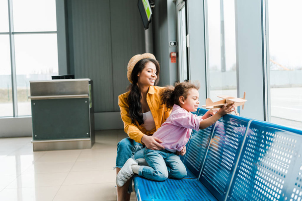 happy african american mother and daughter sitting in airport near window and playing with wooden plane model - 写真・画像