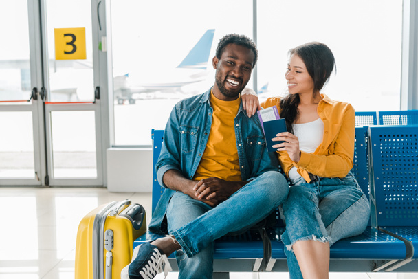 happy african american couple sitting in departure lounge with baggage and air tickets in airport - 写真・画像