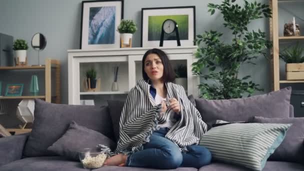 Zoom-in of young woman watching scary thriller on TV sitting on sofa at home - Filmagem, Vídeo
