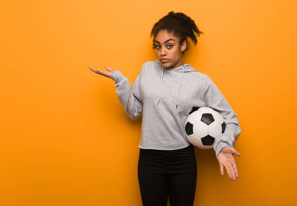 Jonge fitness zwarte vrouw verward en twijfelachtig. Het houden van een voetbal. - Foto, afbeelding