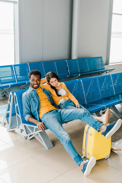 mujer afroamericana agotada y hombre sonriente sentado en la sala de salida con maleta y esperando el vuelo
 - Foto, Imagen