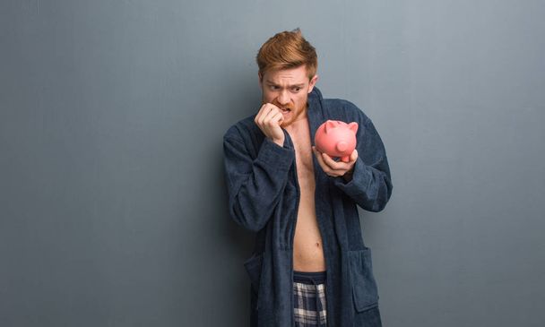 junger rothaariger Mann mit Pyjama, der Nägel beißt, nervös und sehr ängstlich. Er hält ein Sparschwein. - Foto, Bild
