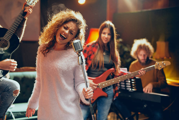 Praktyce Band na show. Kobiety z kręconymi włosami, trzymając mikrofon i śpiewa podczas człowieka w tle gra na gitarze akustycznej. Domowym studio wnętrz. - Zdjęcie, obraz
