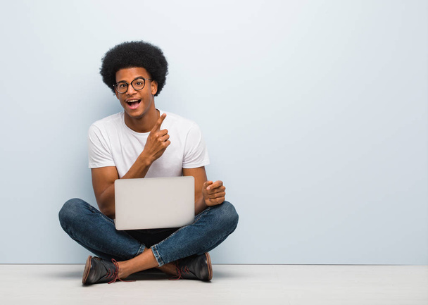 Νέος μαύρος άνθρωπος κάθεται στο πάτωμα με ένα φορητό υπολογιστή που οδηγεί στο πλάι με το δάχτυλο - Φωτογραφία, εικόνα