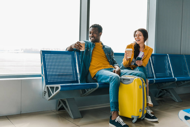 出発ラウンジに座っている幸せなアフリカ系アメリカ人のカップルと黄色のトラベルバッグ - 写真・画像