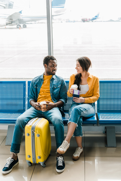 sonriente pareja afroamericana sentada en salón de salida con café para llevar y maleta amarilla
 - Foto, imagen