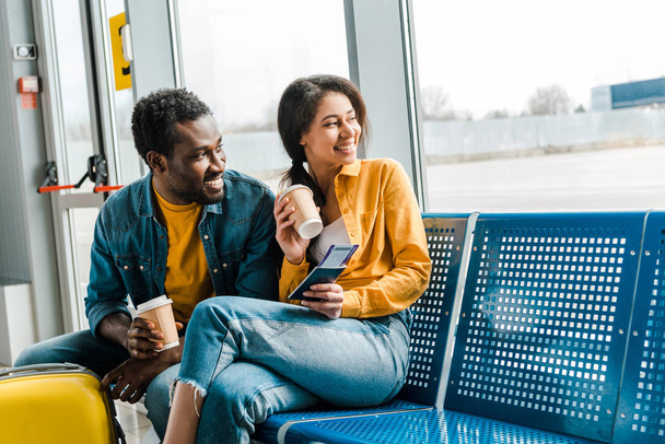 出発ラウンジに座っている幸せなアフリカ系アメリカ人のカップルは、コーヒーを飲みに行き、航空券と窓を見て - 写真・画像