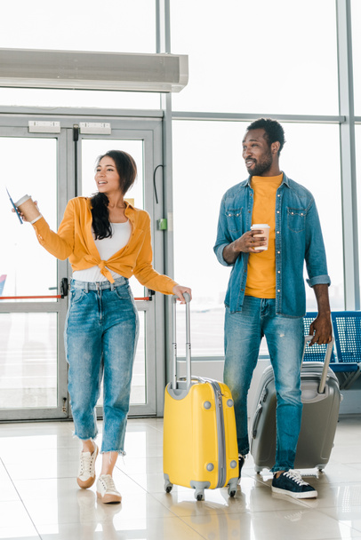 feliz pareja afroamericana caminando juntos en el aeropuerto con bolsas de viaje y café para llevar
 - Foto, Imagen